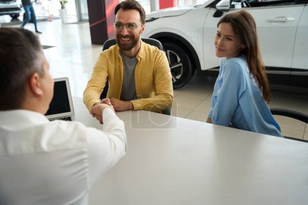 Foto de Married couple communicates with a consultant in the office area of a car dealership, the men greet each other - Imagen libre de derechos