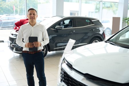 Photo for Employees of a car dealership in office clothes stand by a white car, holding a tablet in their hands - Royalty Free Image