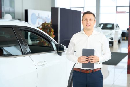 Foto de Man in office clothes stands near a white car in a car dealership, he has a tablet in his hands - Imagen libre de derechos