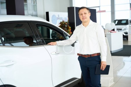 Foto de Customer service manager in office clothes stands by a white car, he works in a car dealership - Imagen libre de derechos