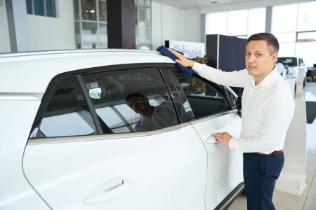 Téléchargez les photos : Man in office clothes wipes the body of a car, he works in a car dealership - en image libre de droit