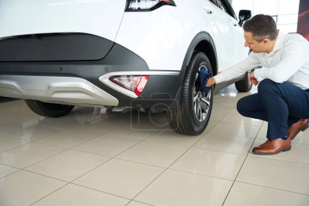 Téléchargez les photos : Man in office clothes wipes the wheels of a car, he works in a car dealership - en image libre de droit