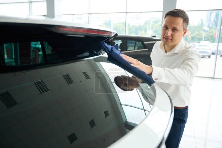 Foto de Man wipes glass of car with a soft napkin, there is a large selection of cars at the car dealership - Imagen libre de derechos