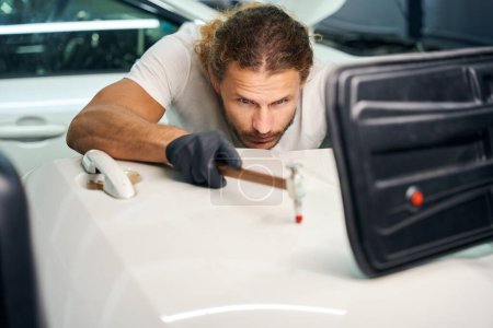 Foto de Guy smoothes out dents on door of a white car, he uses a special straightening tool and a powerful lamp - Imagen libre de derechos