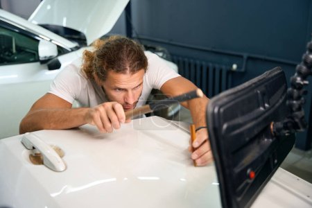 Téléchargez les photos : Auto mechanic straightens the body of a white car with a straightening stick, he works in a car repair shop - en image libre de droit