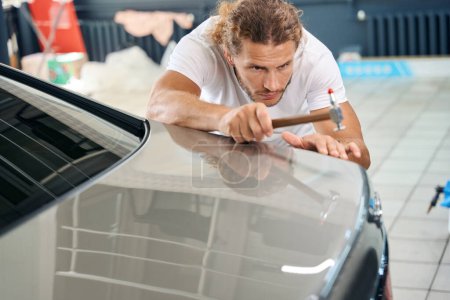 Foto de Experienced master straightens the body of a white car with a straightening hammer, he works in a car repair shop - Imagen libre de derechos