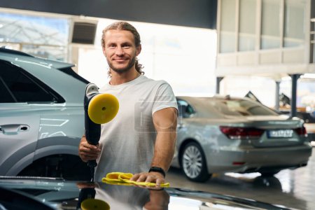 Téléchargez les photos : Smiling guy works in a car repair shop, he polishes a car with a special grinder and a soft napkin - en image libre de droit