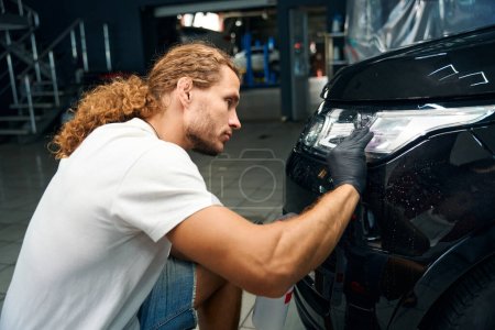 Téléchargez les photos : Guy works in a car repair shop detailing a black car, he uses protective gloves - en image libre de droit