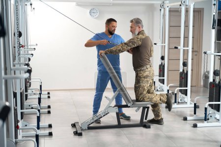 Photo for Patient exercises on a special simulator during the recovery period, he is assisted by an experienced trainer - Royalty Free Image