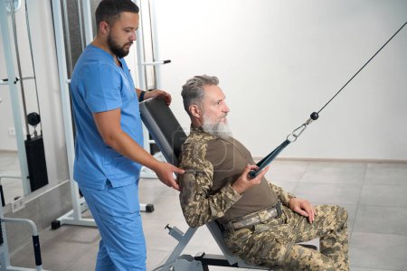 Foto de Man is working out under the guidance of experienced trainer on a special simulator, in a room with modern equipment - Imagen libre de derechos