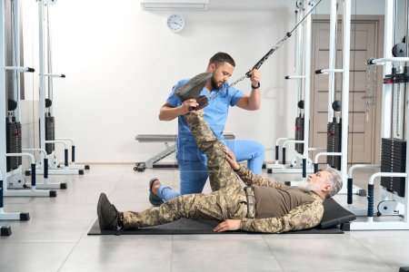 Foto de Specialist physiotherapist helps a military patient with rehabilitation, classes are held on a special simulator - Imagen libre de derechos