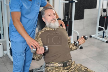 Téléchargez les photos : Bearded man is working out with dumbbells under the guidance of physiotherapist, he is restoring his health in rehabilitation center - en image libre de droit