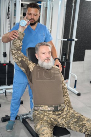 Foto de Bearded man is restoring his health in a rehabilitation center, an experienced physiotherapist is working with him - Imagen libre de derechos
