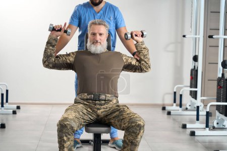 Téléchargez les photos : Bearded man is working out in the gym in a rehabilitation center, an experienced physiotherapist is working with him - en image libre de droit