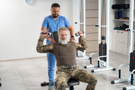 Photo for Man in camouflage clothes works out in the gym in a rehabilitation center, an experienced physiotherapist works with him - Royalty Free Image