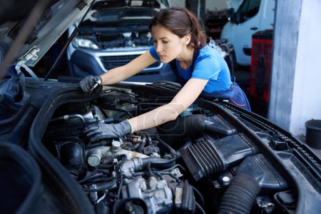 Foto de Encantadora joven mecánico inspecciona el motor bajo el capó de un coche, ella utiliza una herramienta especial - Imagen libre de derechos