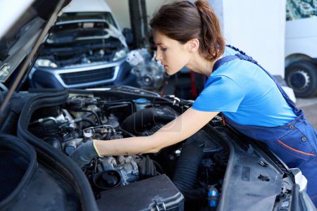 Téléchargez les photos : Mignonne mécanicienne inspecte le moteur sous le capot d'une voiture, elle travaille dans un atelier de réparation - en image libre de droit