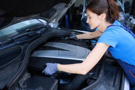 Foto de Auto mecánico trabaja bajo el capó de un coche moderno, una mujer en un uniforme azul - Imagen libre de derechos