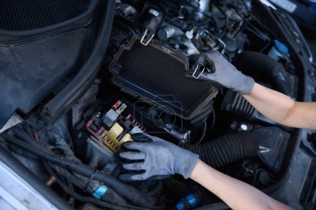 Foto de Taller de reparación de coches mujer comprueba la batería bajo el capó de un coche, el mecánico trabaja en guantes de protección - Imagen libre de derechos