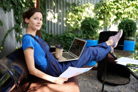 Téléchargez les photos : Une femme souriante s'assoit avec un café et un ordinateur portable dans un coin vert, une femme en uniforme bleu - en image libre de droit