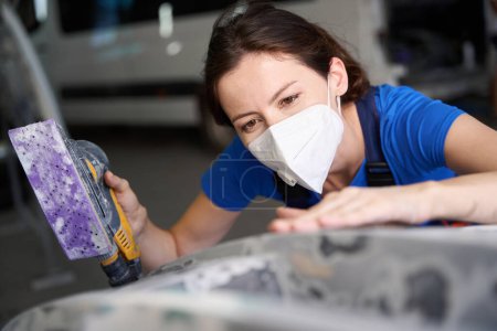 Photo for Auto mechanic in a blue overalls prepares a car for painting the body, a woman uses a grinding machine - Royalty Free Image
