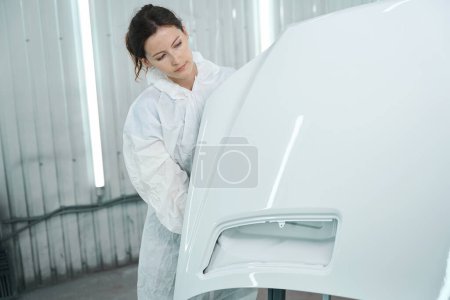 Photo for Craftswoman in white protective equipment works in a paint shop, she has a white car part - Royalty Free Image