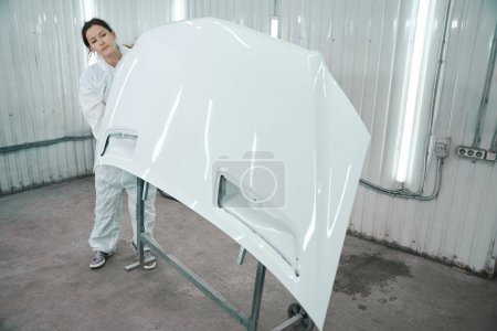 Téléchargez les photos : Femme en tenue de protection blanche travaille dans un atelier de peinture, elle a une pièce de voiture blanche - en image libre de droit