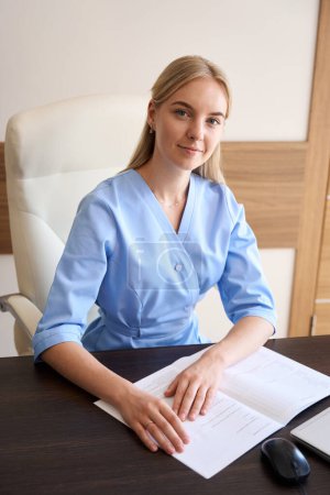 Foto de Joven doctora o enfermera europea sentada en el escritorio con copybook abierto y mirando a la cámara. Concepto sanitario - Imagen libre de derechos