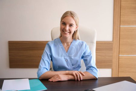 Photo for Front view of young smiling caucasian female doctor sitting at working desk with documents and laptop and looking at camera. Healthcare concept - Royalty Free Image
