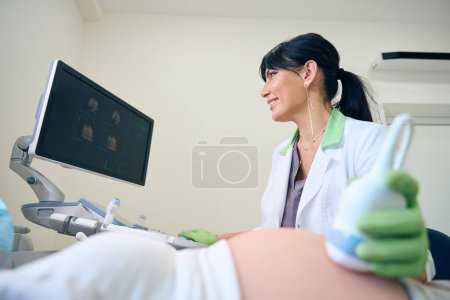 Foto de Ginecóloga europea adulta observando en monitor durante el examen por ultrasonido de una mujer embarazada parcial en la clínica. Concepto de examen de embarazo - Imagen libre de derechos