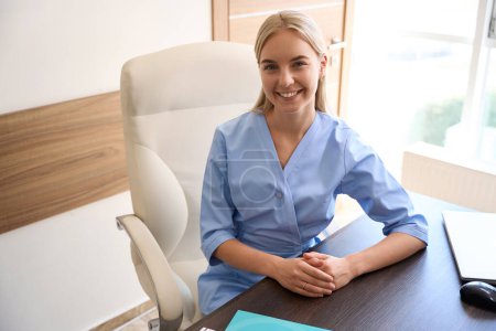 Foto de Joven mujer caucásica sonriente médico o enfermera sentada en el escritorio con documentos y portátil y mirando a la cámara. Concepto sanitario - Imagen libre de derechos