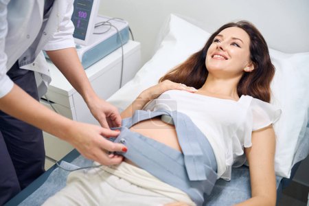 Foto de Ginecóloga femenina recortada preparando vientre de joven sonriente mujer embarazada caucásica para ecg en clínica. Concepto de examen de embarazo - Imagen libre de derechos