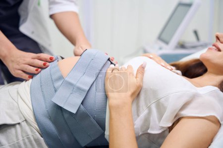 Téléchargez les photos : Gynécologue féminine partielle préparant le ventre de la femme enceinte souriante pour ecg en clinique. Concept d'examen de grossesse - en image libre de droit