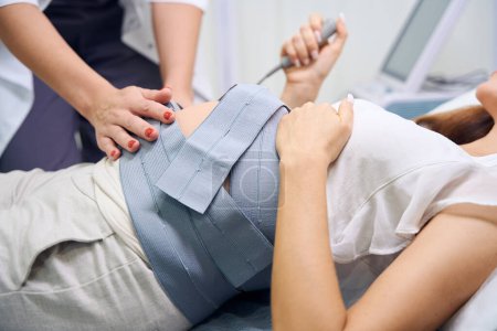 Téléchargez les photos : Image recadrée de gynécologue féminine préparant le ventre de la femme enceinte pour ecg en clinique. Concept d'examen de grossesse - en image libre de droit