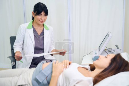 Foto de Ginecóloga europea adulta sosteniendo electrocardiograma de vientre de mujer embarazada caucásica joven y mirándola en clínica. Concepto de examen de embarazo - Imagen libre de derechos