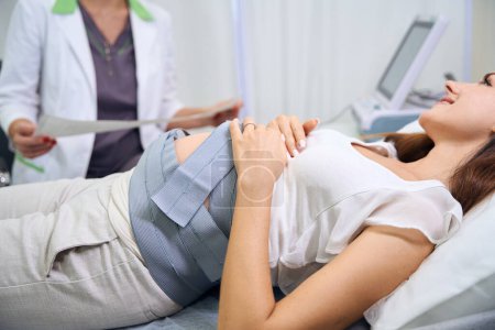 Photo for Blurred partial female gynecologist checking electrocardiogram of belly of young caucasian pregnant woman in clinic. Concept of pregnancy examination - Royalty Free Image