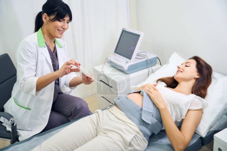 Foto de Ginecóloga europea adulta mostrando a la joven mujer embarazada caucásica electrocardiograma de su vientre en la clínica. Concepto de examen de embarazo - Imagen libre de derechos