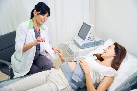 Photo for Adult european female gynecologist showing to young smiling caucasian pregnant woman electrocardiogram of her belly in clinic. Concept of pregnancy examination - Royalty Free Image
