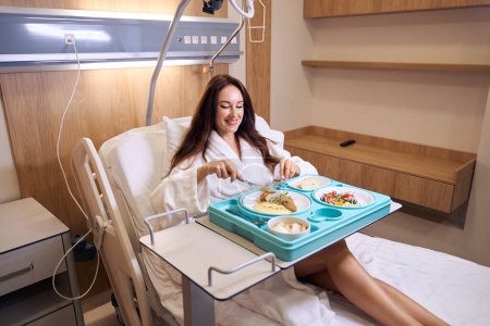 Photo for Young smiling caucasian pregnant woman wearing bathrobe cutting meat with knife and fork during having lunch on medical couch in clinic room. Concept of pregnancy and maternity - Royalty Free Image