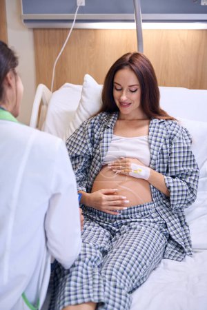 Téléchargez les photos : Recadrage de l'infirmière examinant la jeune femme enceinte caucasienne avant l'accouchement sur le lit médical à l'hôpital. Concept de grossesse et de maternité - en image libre de droit