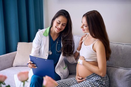 Foto de Enfermera europea sonriente y joven mujer embarazada caucásica mostrando algo en el portapapeles antes del parto en el sofá en la sala médica de la clínica. Concepto de embarazo y maternidad - Imagen libre de derechos
