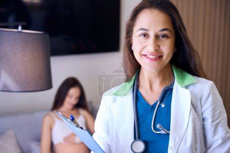 Photo for Portrait of young smiling european female nurse looking at camera with blurred pregnant woman on background in medical room of clinic. Concept of pregnancy and maternity - Royalty Free Image
