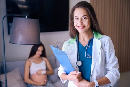 Foto de Enfoque selectivo de la joven enfermera europea sonriente mirando a la cámara con el fondo de la mujer embarazada borrosa en el sofá en la sala médica de la clínica. Concepto de embarazo y maternidad - Imagen libre de derechos