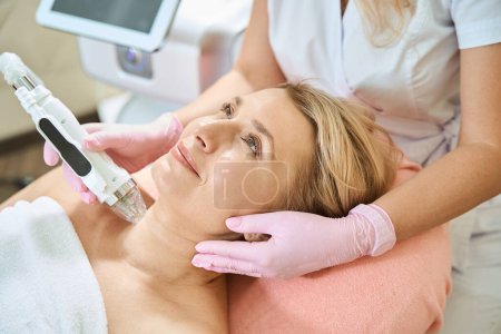 Photo for Cropped female cosmetologist doing microneedle rf lifting of neck skin with IPL photorejuvenation device of smiling adult caucasian woman in blurred beauty salon. Body skin care and rejuvenation - Royalty Free Image