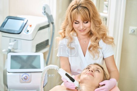 Photo for Female european beautician doing microneedle rf lifting of neck skin with IPL photorejuvenation device of smiling adult caucasian woman in blurred beauty salon. Body skin care and rejuvenation - Royalty Free Image