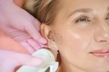 Photo for Close up of cropped female cosmetologist doing microneedle rf lifting of ear skin with IPL photorejuvenation device of smiling adult caucasian woman. Concept of body skin care and rejuvenation - Royalty Free Image