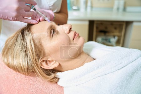 Foto de Vista lateral de una cosmetóloga parcial haciendo inyección de belleza facial con jeringa de mujer caucásica adulta en un salón de belleza borroso. Cuidado, elevación y rejuvenecimiento de la piel facial - Imagen libre de derechos
