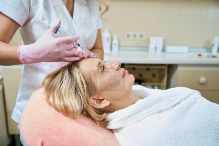 Photo for Side view of partial female cosmetologist doing face beauty injection with syringe of adult caucasian woman wearing bathrobe in blurred beauty salon. Face skin care, lifting and rejuvenation - Royalty Free Image