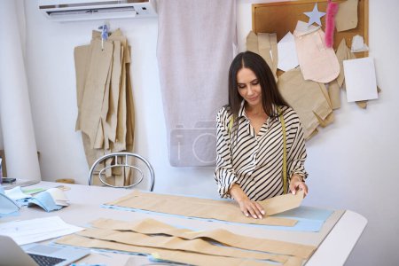 Téléchargez les photos : Jeune brune à une table de découpe pose un motif sur le tissu, une femme dans un chemisier à rayures confortable - en image libre de droit