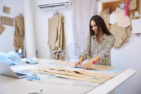 Foto de Sonriente morena en una mesa de cortar establece un patrón en la tela, una mujer en una cómoda blusa a rayas - Imagen libre de derechos
